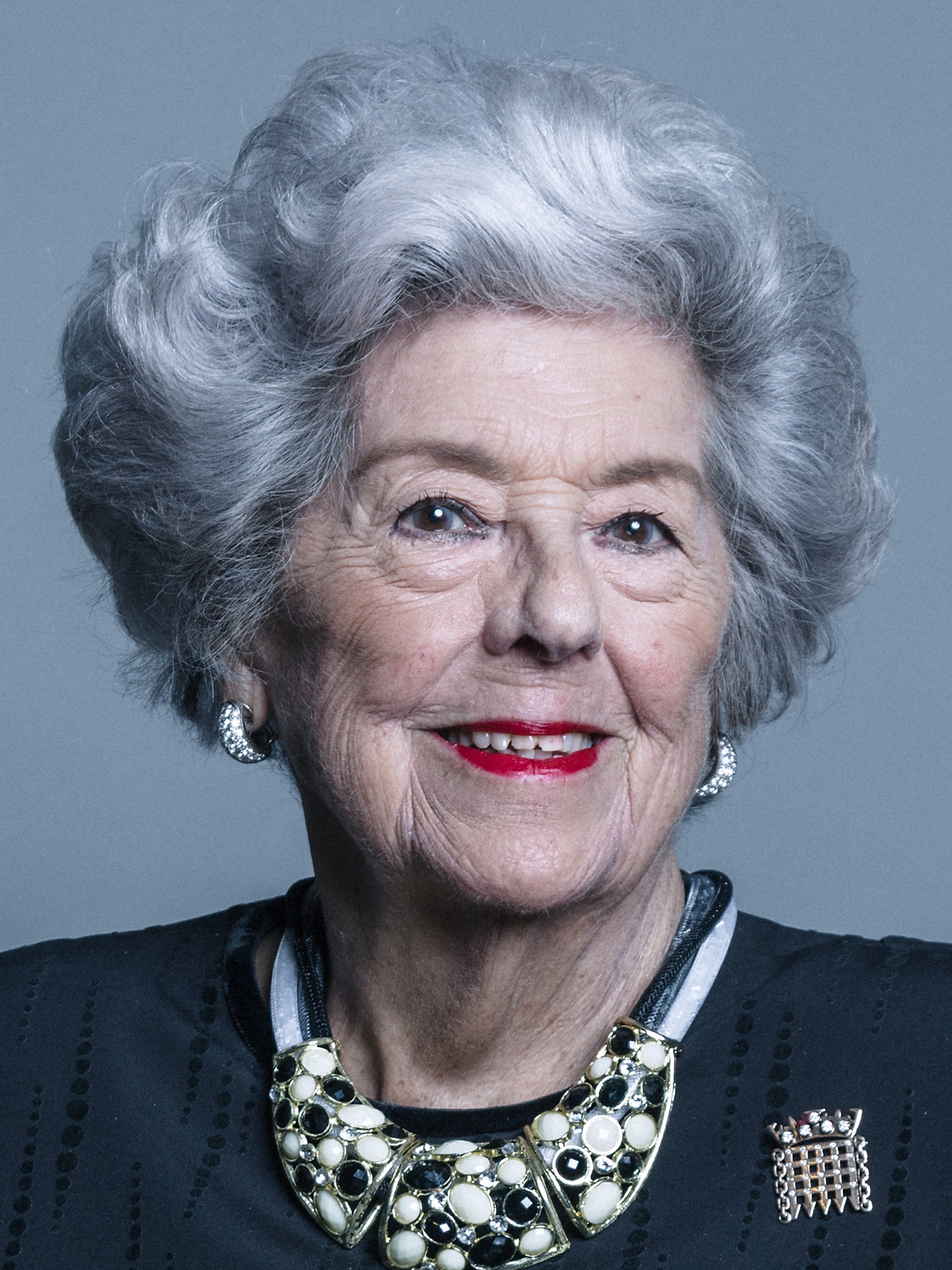 Photograph of a white woman's shoulders and face. She has light eyes, wrinkles, grey short styled hair. She is wearing red lipstick, earrings, a large necklace decorated with black and white gems, a portcullis broach. The background is grey.